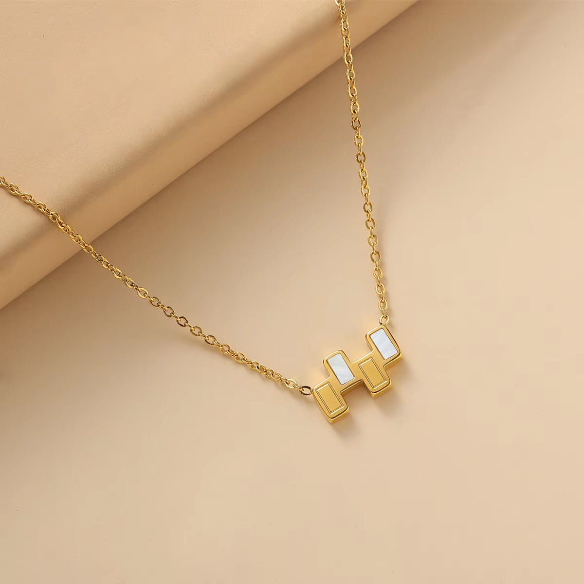 A gold chain necklace with a geometric pendant featuring white inlay details, displayed on a beige background.