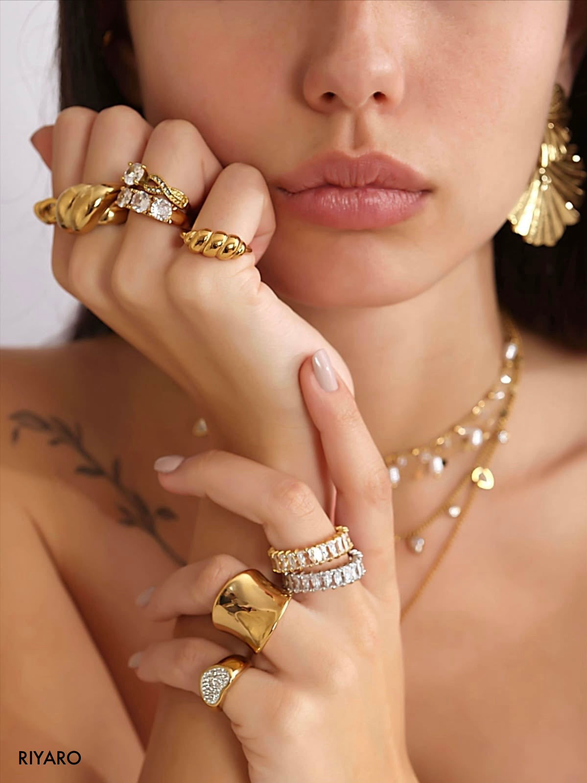 A close-up lifestyle shot of a model wearing multiple gold rings, some with clear stones, layered with gold necklaces and earrings. The brand name 'RIYARO' is visible in the bottom-left corner.