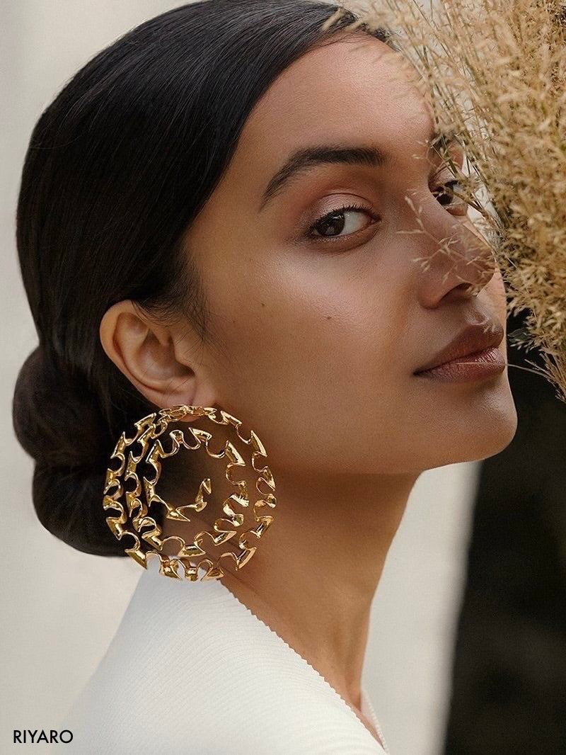 A close-up lifestyle shot of a model wearing a pair of large, chunky gold chain-link hoop earrings. The model has dark hair styled in a low bun and is wearing a white top, with dried grass elements in the background.