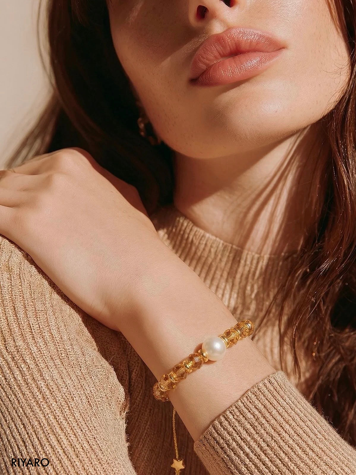 A close-up lifestyle shot of a model wearing a gold chain bracelet with a white pearl and a dangling gold star charm, paired with a beige ribbed knit sweater.