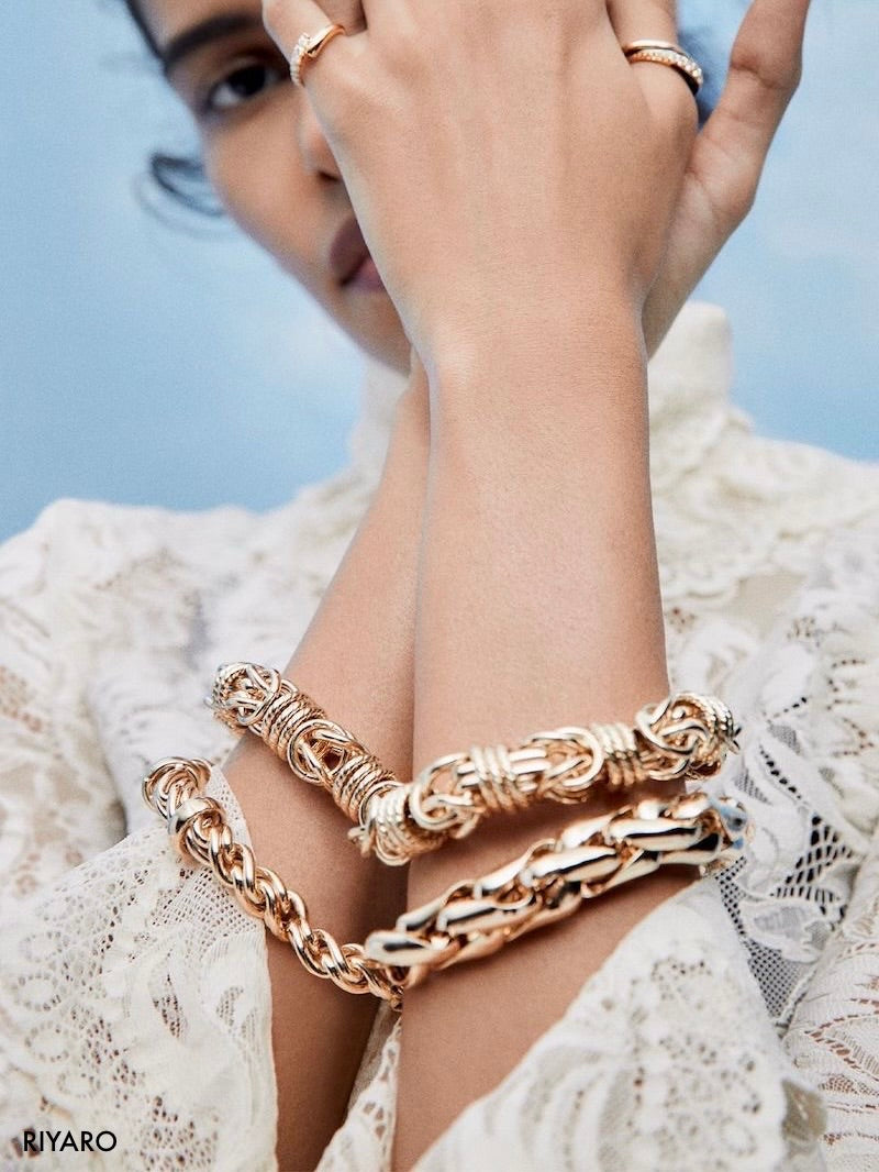 A close-up lifestyle shot of a model wearing multiple chunky gold chain and textured link bracelets on her wrist, along with matching gold rings on her fingers. She is wearing a white lace top against a soft blue sky background.