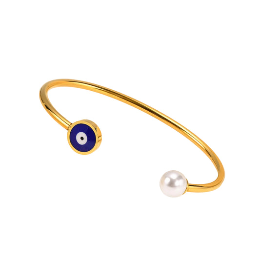 Gold-tone open cuff bracelet with a blue evil eye charm and a white pearl bead, displayed on a cream-colored jewelry stand.
