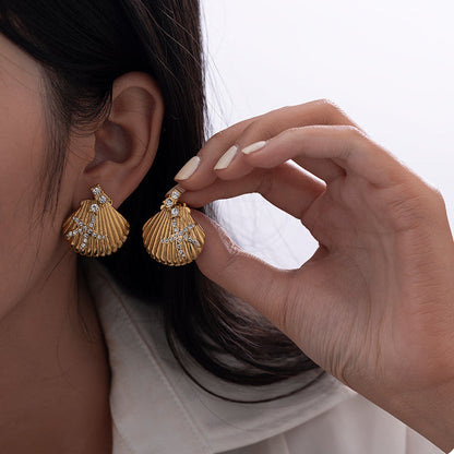 A close-up lifestyle shot of a model wearing gold shell-shaped dangle earrings with clear rhinestone star accents, with one earring on her ear and the other held by her hand.